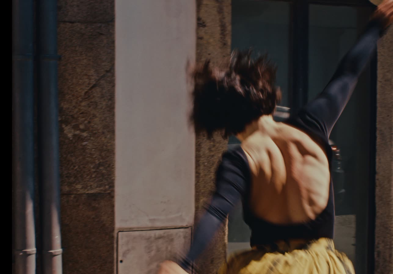 Dancer standing between two clotheslines of hanging dresses in an outdoor alley, Em Quanto Espero