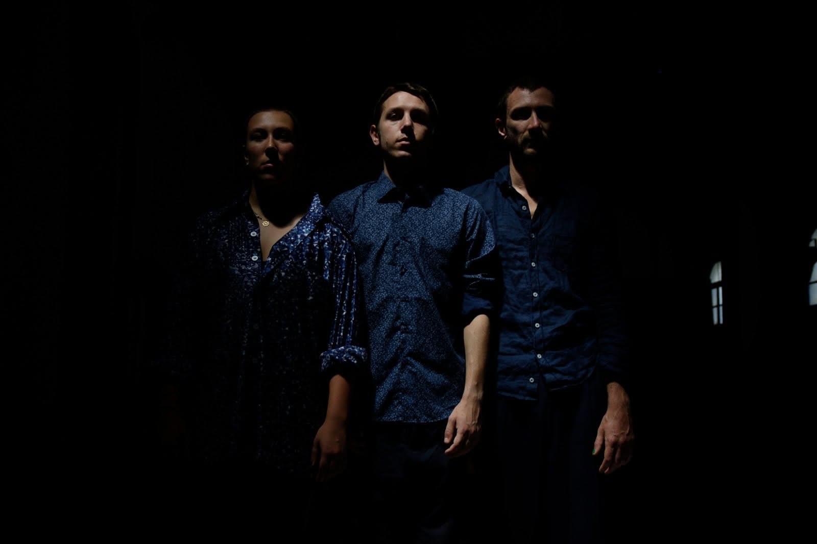 Three performers in blue shirts standing in dim light, Everyours