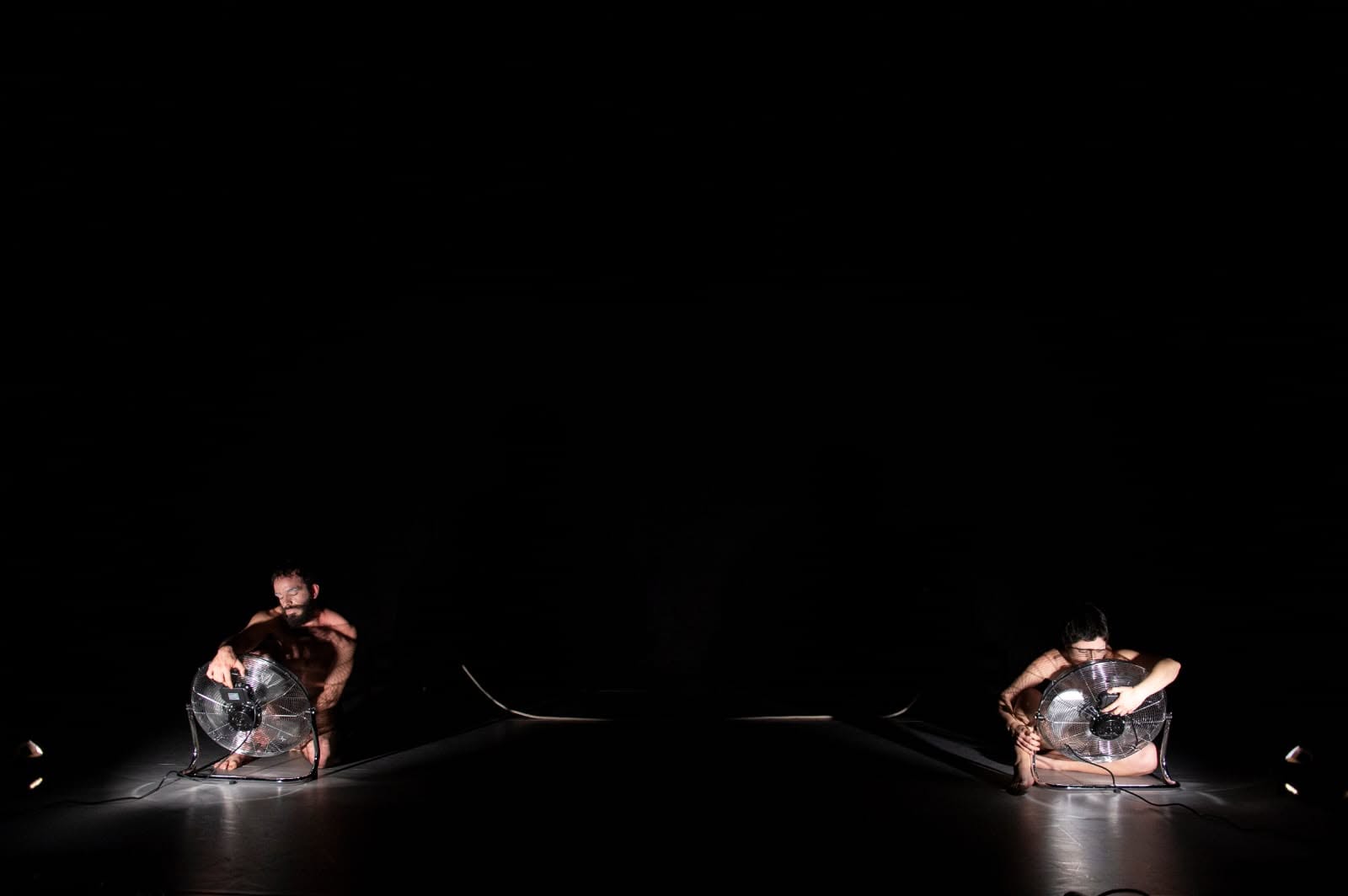 Two performers kneeling with electric fans on a dark stage, In the Absence of Tenderness